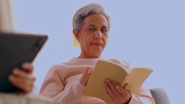 Senior woman with gray hair reading book near tablet at home enjoying technology leisure indoors calm relaxing couch quiet room paper novel digital device
