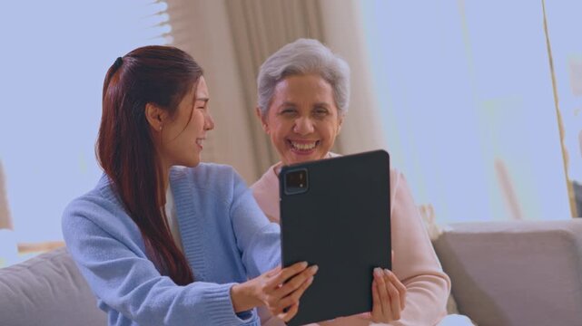Asian woman senior selfie tablet smile home family couch living room, cheerful daughter elderly mother taking vdo together indoors cozy sofa generation