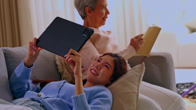 Senior woman with tablet on couch reading book at home living room, warm smile, relaxed leisure, cozy family bonding, digital device and quiet indoor moment