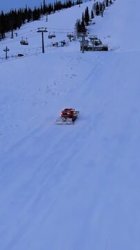 Red snow groomer with lights on compacting and leveling the snow on a mountain slope at dusk, preparing the piste for the next day of skiing at a winter resort with a chairlift
