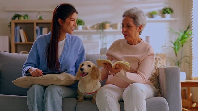 Young woman with elderly woman, dog, sofa, book and tablet at home in living room, family reading, conversation, caregiver support, friendship with pet indoors