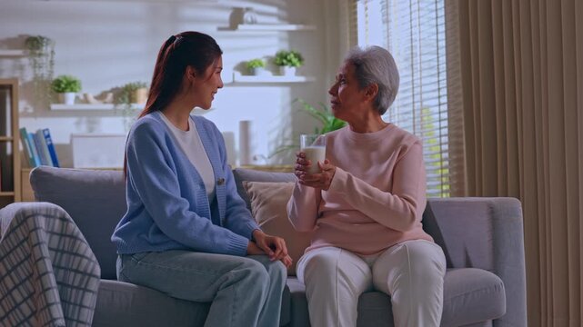Asian senior woman caregiver offering milk on sofa in living room with gentle home care support, warm drink, calm conversation, smile, companionship, comfort, elderly wellness