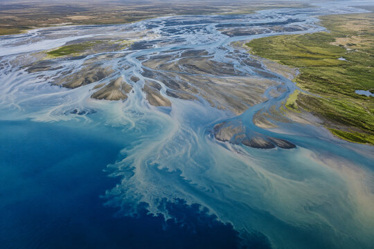 Aerial perspective of braided river system carrying glacial sediment into deep blue ocean forming artistic natural silt patterns and delta textures.