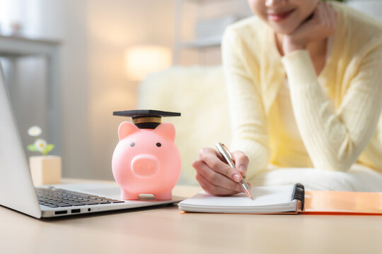 Woman planning education savings with piggy bank in graduation cap, laptop and notebook at home, representing college fund, student finance, budgeting and future education goals.