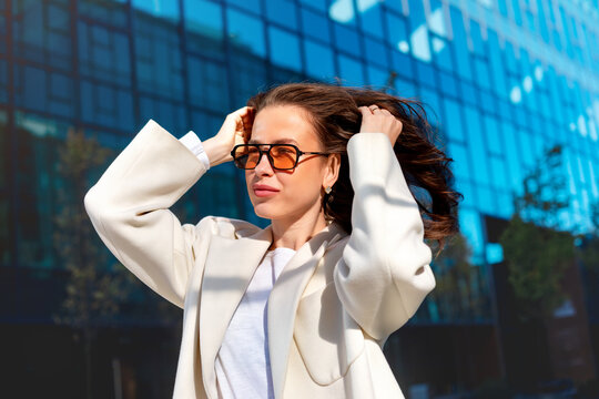 Beautiful woman adjusting windblown hair in elegant white blazer before modern office building. Portrait of female model wearing stylish sunglasses, business style, urban fashion