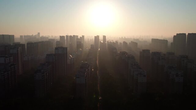 Aerial view of dense urban buildings under soft hazy sunrise light, with bright sun above the city skyline.