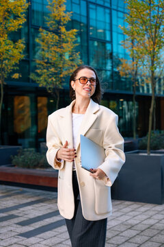 Young woman smiling confidently with laptop in elegant white blazer. Beauty woman presenting executive business style and sophisticated urban fashion outdoors.