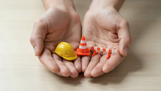 Hands holding miniature construction helmet traffic cone and safety barrier