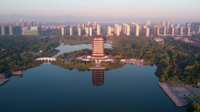 An aerial view shows a traditional pagoda by a lake in a green urban park, with modern city buildings in the distance.