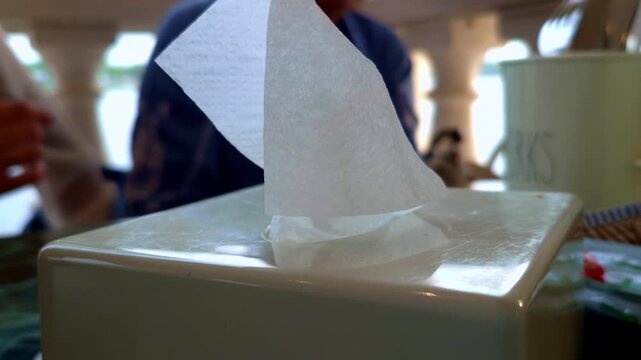 A white tissue protrudes from a rectangular tissue box in close-up view. A softly blurred person and table setting create a simple indoor background.