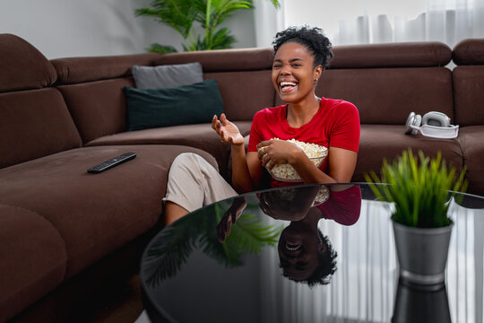Woman enjoying a movie night at home while laughing and eating popcorn on the sofa