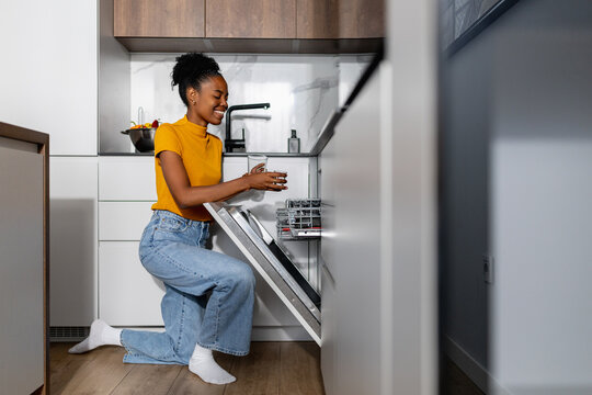 Woman kneeling by an open dishwasher while loading clean dishes in a modern kitchen setting