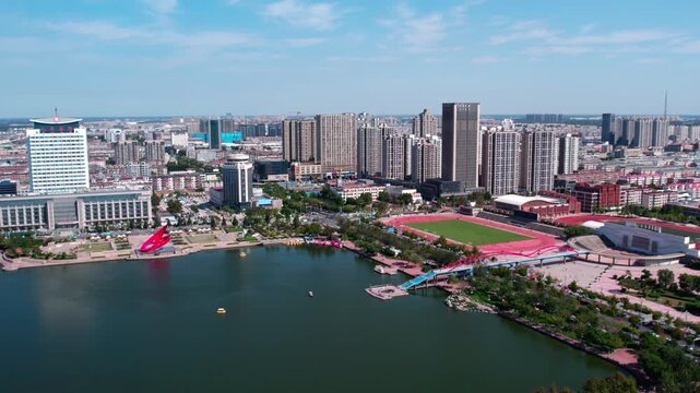 A panoramic view of a modern city featuring a lake, high-rise buildings, and a lakeside sports facility on a clear sunny day.
