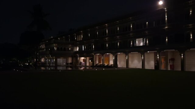 Udaipur's illuminated heritage-style palace features rows of arched balconies and warm lights. A quiet lawn beneath the night sky creates an elegant nighttime atmosphere.