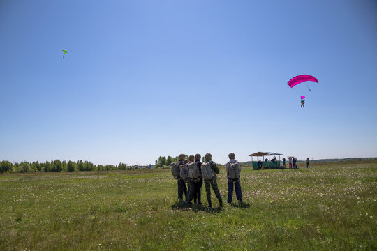Group of skydivers standing in grassy field watching two parachutes descending against clear blue sky, parachuting training session outdoor scene