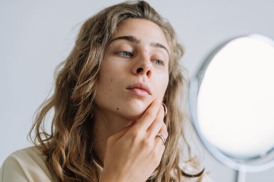 A portrait of a frustrated woman examining her acne-prone skin in a small mirror. A sad woman touches her face, touching her pimples indoors. Concept of skincare, acne, and breakouts.