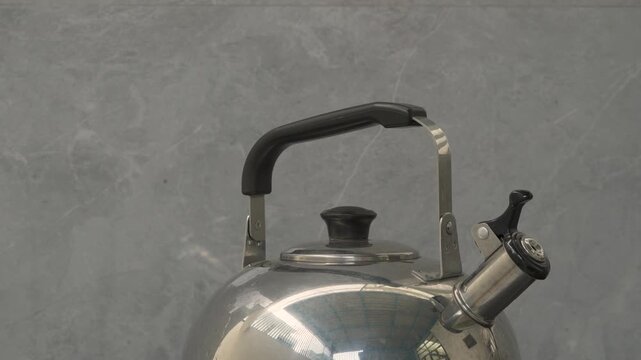 A close-up shot of a stainless steel whistling tea kettle with a black handle and lid knob against a light gray textured background. Steam gently wafts from the spout, indicating boiling water