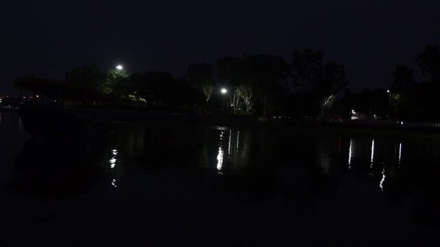 Trees and streetlights line Udaipur's far shore at night, with bright reflections shimmering vertically across the dark water. The quiet scene has a subdued atmosphere.