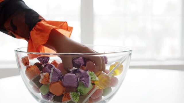 Child hand grabbing colorful wrapped candies from glass bowl during festive fall holiday season celebration with filled assorted colorful orange sleeves reaching
