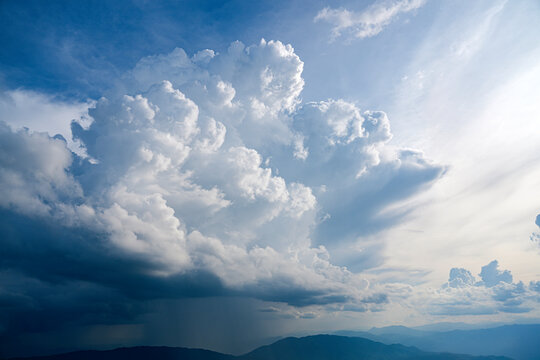 Lush, white clouds swirl and gather in dramatic formations while glimpses of sunlight illuminate the serene landscape below on a calm afternoon