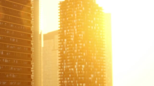 Tall buildings stand in an urban area as sunlight reflects off the glass surfaces. It is late afternoon and the sun is setting, creating bright light and long shadows.