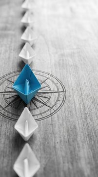 Blue Paper Boat Leading A Fleet Of Small White Boats With Compass Icon On Wooden Table With Sunlight - Leadership Concept