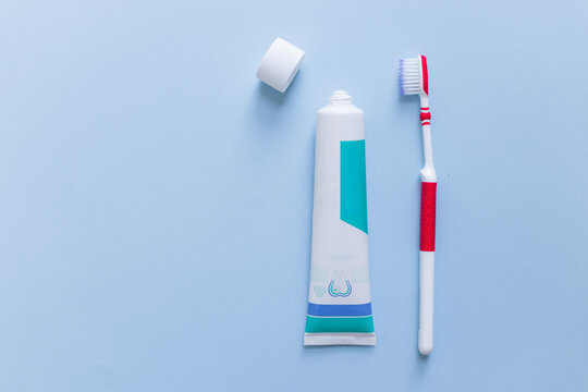 Flat lay of an open toothpaste tube with cap and a red toothbrush side by side on a light blue background. Daily oral hygiene and dental care concept.