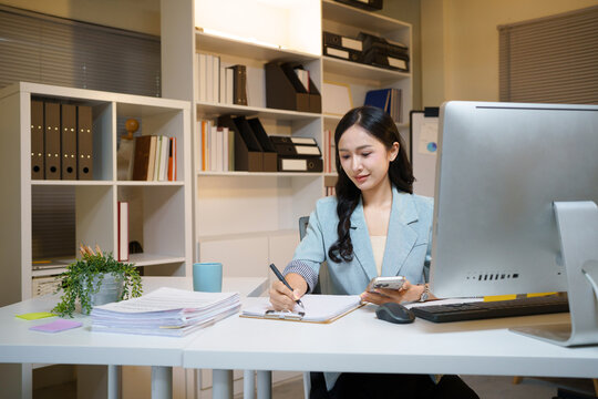 asian businesswoman working at her desk, focused on taking important notes on a notebook while using her smartphone, ideal for business and corporate concepts.