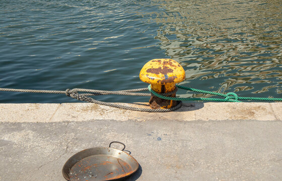 Weathered yellow mooring bollard with nautical rope and empty paella pan on harbor pier