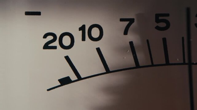 Mechanical indicator needle moving quickly across numbered decibel scale of analog VU volume meter. Macro shot