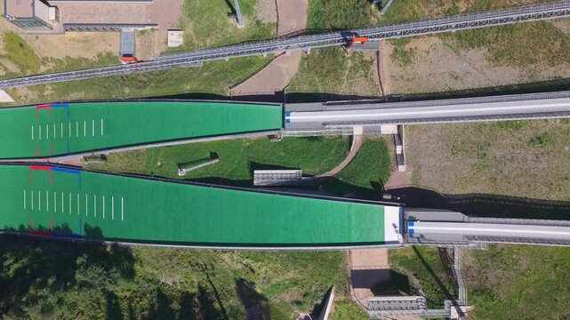 Top-down aerial view of Predazzo Ski Jumping Stadium in the Dolomites, Val di Fiemme, Italy. A geometric summer training facility and Olympic venue surrounded by Alpine forest.