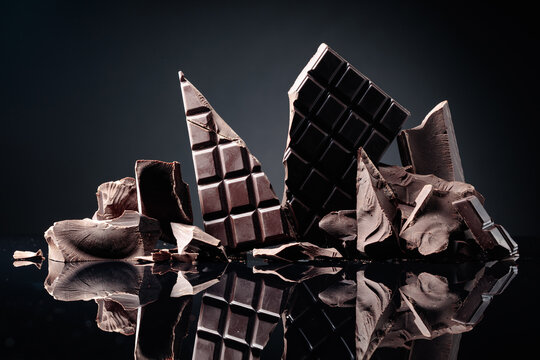 A broken chocolate bar and pieces of dark chocolate on a black reflective background.
