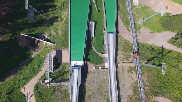 Top-down aerial view of Predazzo Ski Jumping Stadium in the Dolomites, Val di Fiemme, Italy. A geometric summer training facility and Olympic venue surrounded by Alpine forest.
