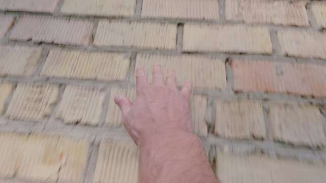 The man carefully feels the brick wall with his hand. The hand gently touches the wall with its fingers. From the first person.