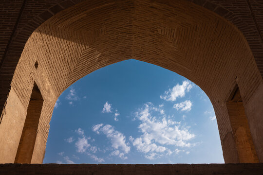 Interior architectural view of the historic Jameh Mosque of Ardestan in Iran, featuring a monumental brick iwan arch framing a vivid blue sky with scattered white clouds.