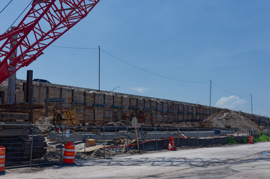 Active road construction at an underpass bridge showing concrete supports, fencing, orange safety barrels and equipment, conveying repair work, urban infrastructure maintenance and site progress.
