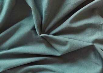 Background view of softly draped teal blue-green fabric with natural folds and gathered texture, creating an elegant abstract textile backdrop with subtle shadows, rich depth, and dimensional detail. © Antonios