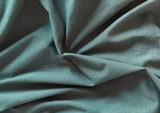 Background view of softly draped teal blue-green fabric with natural folds and gathered texture, creating an elegant abstract textile backdrop with subtle shadows, rich depth, and dimensional detail.