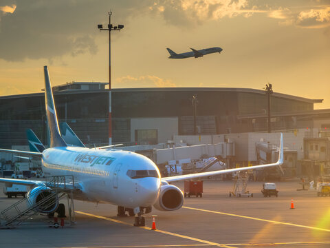 Calgary, AB, Canada. August 16, 2026. WestJet Boeing airplane parked at an airport gate during golden hour sunset while another jet departs.