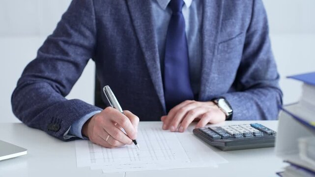 Businessman wearing suit and tie is using calculator and taking notes while working at desk in office, with laptop and stack of folders nearby, close up. Audit and taxes theme in business