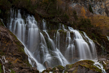 Fototapeta premium Silky cascading water flows over mossy rocks beneath autumn foliage in the Swiss countryside.