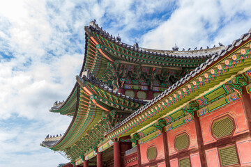 Obraz premium A low-angle view shows colorful painted eaves and tiled roofs on a traditional Korean wooden building beneath a cloud-filled blue sky.
