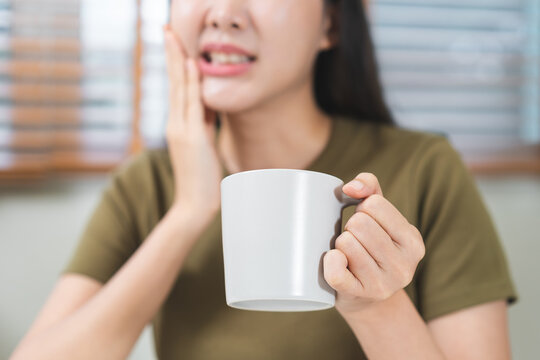 Asian young woman holding a mug and touching her cheek in pain, suffering from sensitive teeth or toothache problem while drinking hot coffee at home, dental healthcare concep.