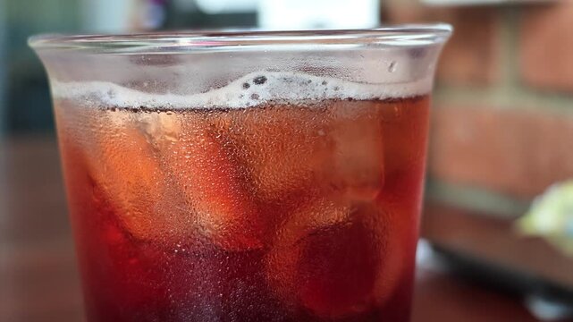 Side view close-up of cold iced lemon tea in a sweaty glass covered with water condensation, showing floating ice cubes and a rustic red brick background.
