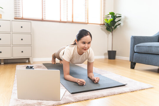Asian young woman practicing yoga in a forearm plank pose while watching an online fitness training tutorial on a laptop at home, core strength workout and digital wellness concept.