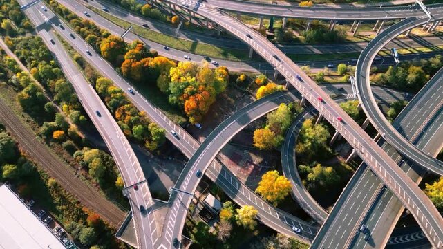 Aerial view of complex highway interchange with moving cars at sunset showing dynamic traffic flow across bridge architecture and autumn forest landscapes.