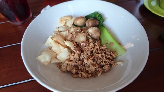 High angle view showing a full ceramic bowl filled with Indonesian chicken noodles, crisp wonton crackers, seasoned minced meat, braised quail eggs, and bok choy.

