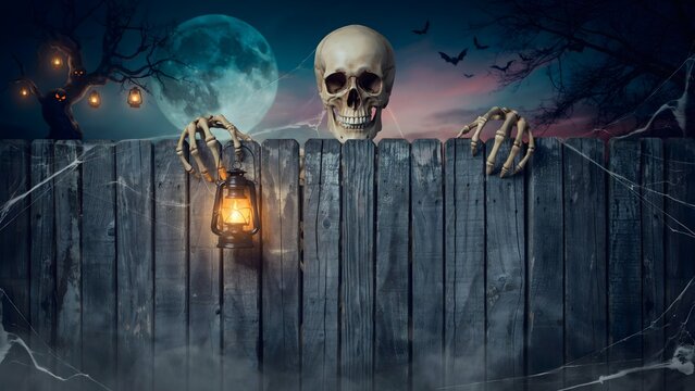 Scary human skull holding a vintage lantern behind a wooden fence at night for Halloween