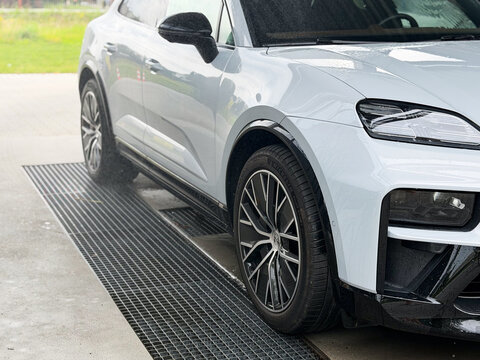 Close-up of a white suv parked on a metal grate in a wet car wash bay, showing front wheel, body side and headlight porsche macan 4 electric Poland, Pszczyna, 3.08.2026
