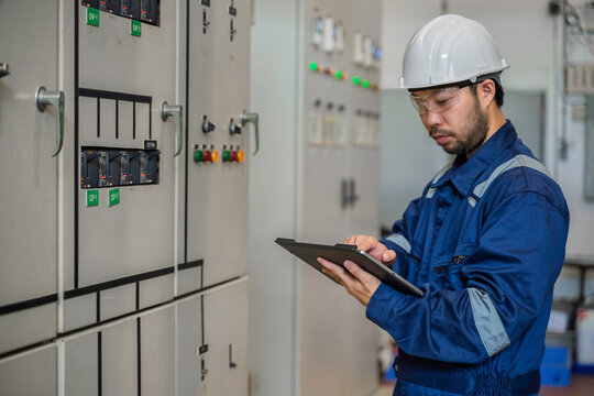 Focused professional male industrial electrical engineer safely uses a digital tablet to inspect a large power control panel during maintenance inside a modern factory manufacturing plant.